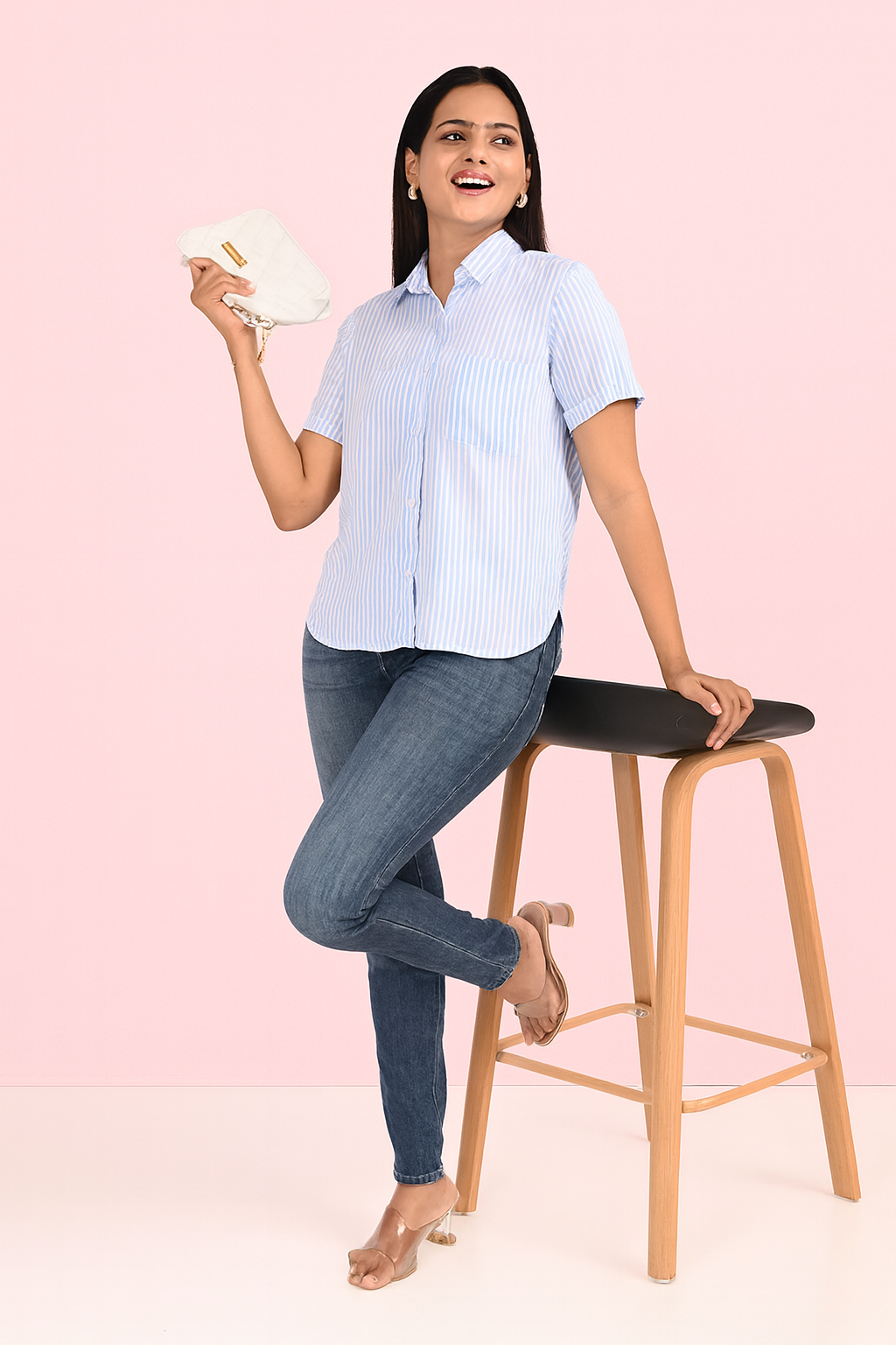 “Woman wearing light blue striped shirt and blue jeans, sitting on a wooden stool holding a white clutch – modern casual look by Classy Clothing.”